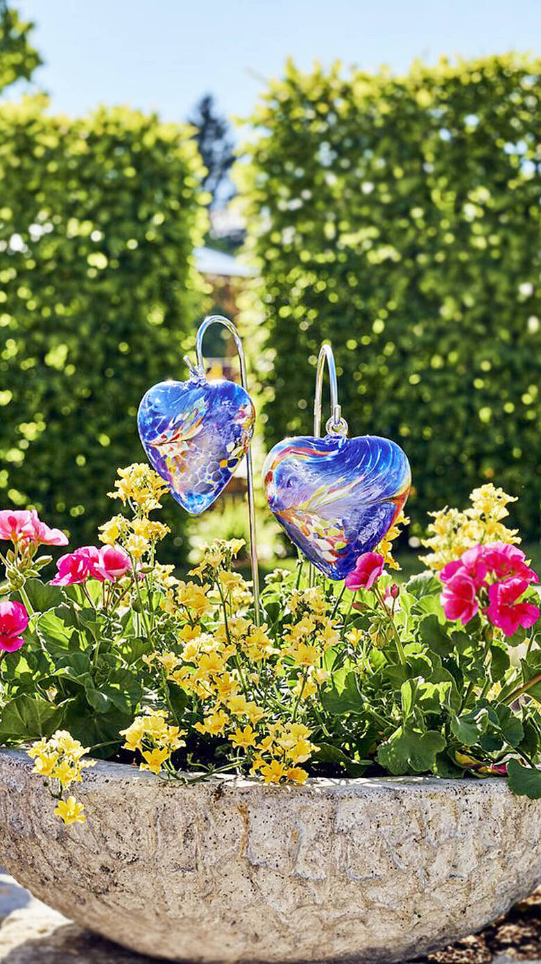 Farbenfrohe Glasherzen in Steinblumenschale zwischen Frühlingsblumen im Garten des JOSKA Glasparadies