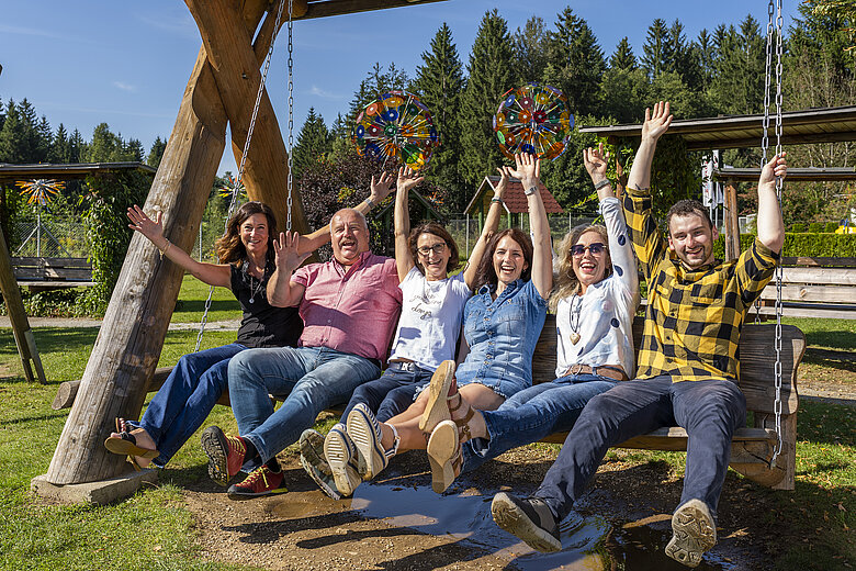 Fröhliches Team beim JOSKA Glasparadies in Bodenmais schaukelt gemeinsam auf einer großen Holzschaukel und hebt lachend die Arme – Symbolbild für Teamgeist und Jobangebote.